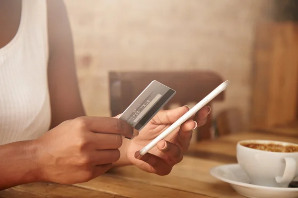Selective focus. Close up shot of female hands holding plastic card and cell phone. African woman paying with credit card while shopping online, using mobile phone, having cappuccino at a restaurant — стоковое фото