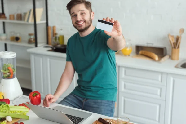 Улыбающийся Человек Показаны Кредитной Карты Столом Ноутбуком Кухне Дома — стоковое фото